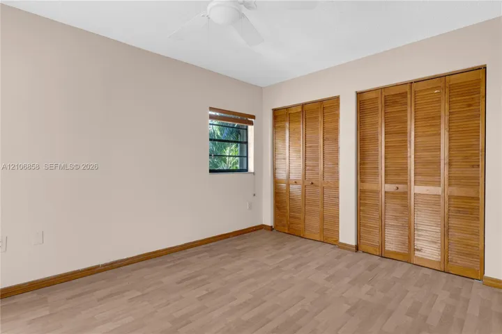 Interior space featuring wood-finish flooring, two sets of bifold wooden closet doors, a ceiling fan with integrated lighting, and a window with wood-slat blinds