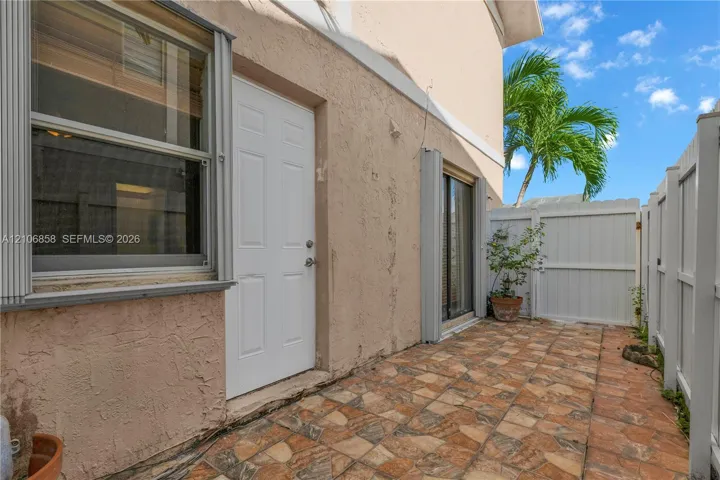 Stucco exterior with multi-pane window, white paneled entry door, sliding glass door, and tiled patio enclosed by white vinyl fencing