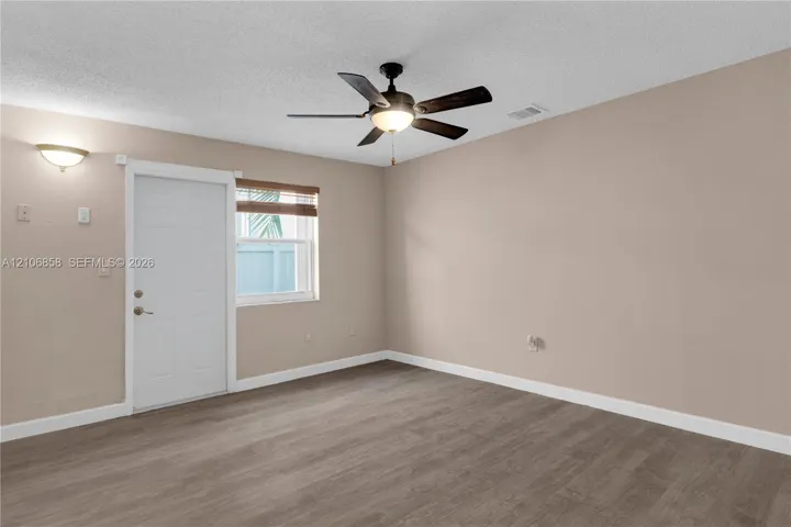 Neutral-toned interior room featuring wood-finish flooring, a ceiling fan with integrated lighting, a single exterior door, and a wall-mounted sconce