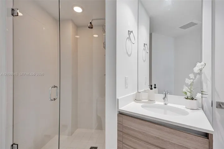 Bathroom featuring a wood-finish vanity with a white countertop and oval undermount sink, large wall-mounted mirror, and glass-enclosed shower with tiled walls