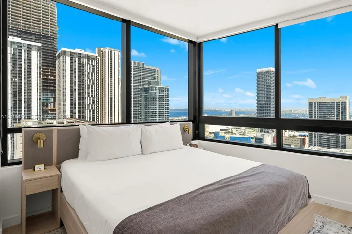 Floor-to-ceiling windows featuring panoramic city and water scenery, light wood-finish built-in headboard with integrated nightstands, and metallic wall-mounted sconces