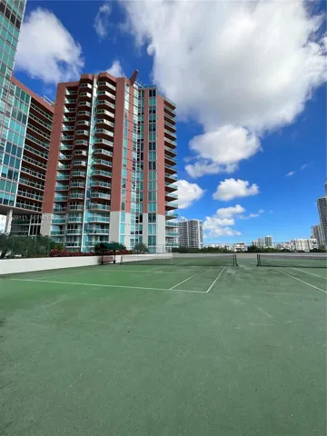 Tennis court with a green hard surface and white court markings, enclosed by chain-link fencing, situated on an outdoor elevated deck