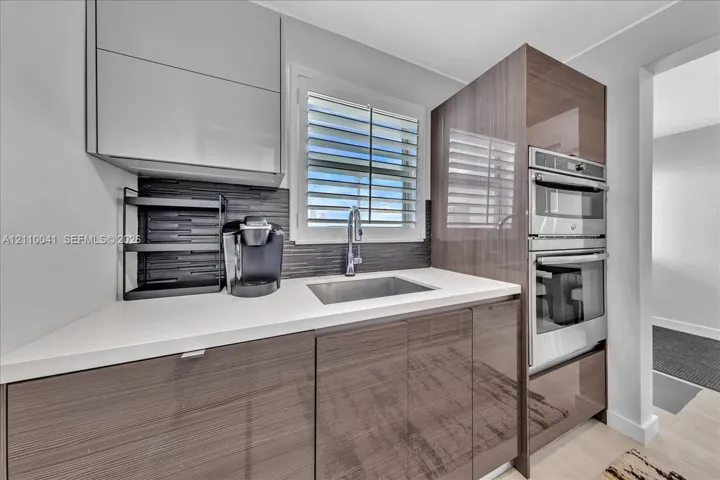 High-gloss cabinetry, stone-finish countertops, stainless steel dual wall oven, dark linear tile backsplash, and undermount stainless steel sink