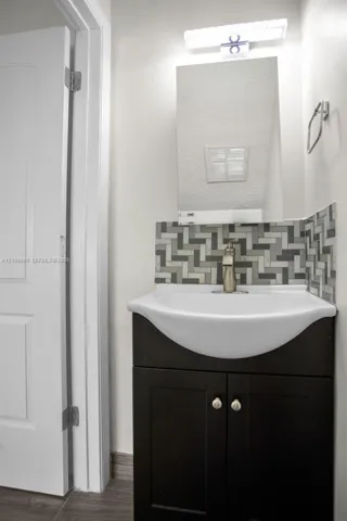 Dark wood-finish vanity with a white integrated basin, multi-tonal herringbone tile backsplash, metallic faucet, wall-mounted rectangular mirror, and bar-style light fixture