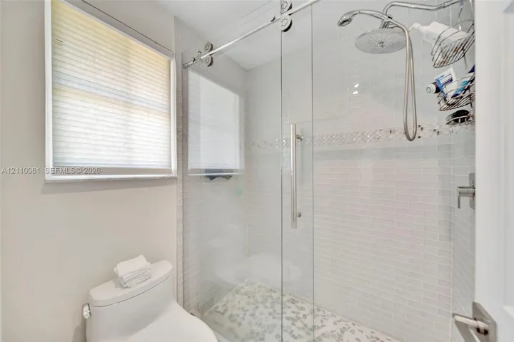 Walk-in shower featuring a glass sliding door, polished metal rain showerhead with handheld attachment, horizontal white tile walls with a decorative mosaic inlay, and textured stone floor tiling