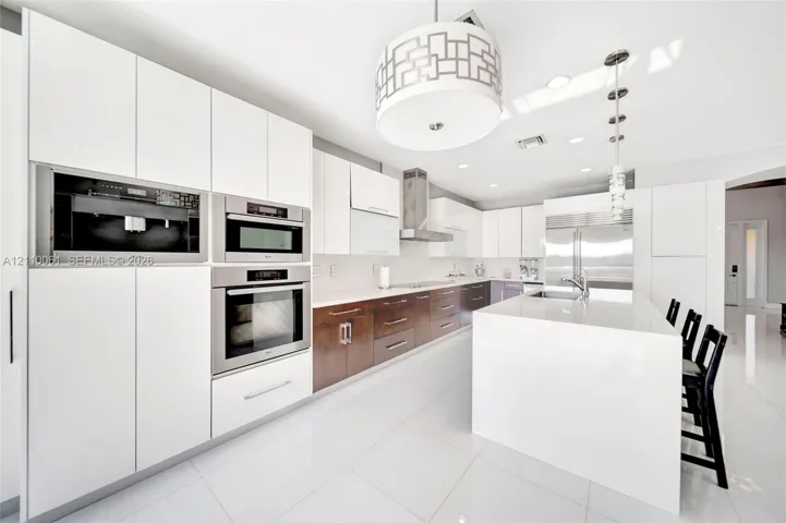 Kitchen featuring two-tone flat-panel cabinetry, a central waterfall-edge island, stainless steel built-in double wall ovens, a coordinating microwave, and a geometric-patterned drum pendant light