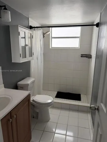 Bathroom featuring white tile flooring, a shower with white wall tile, a wood-finish vanity with a sink, and a white wall-mounted storage cabinet