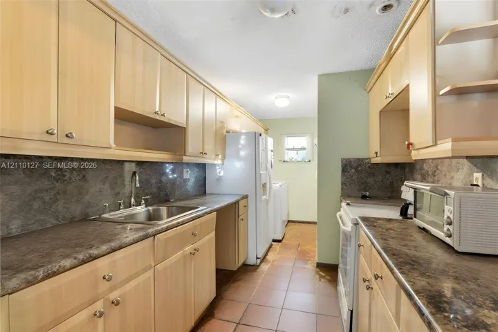 Kitchen featuring wood-finish cabinetry, stone-texture laminate countertops, dark stone-style backsplash, integrated stainless steel sink with metal faucet, and tile flooring