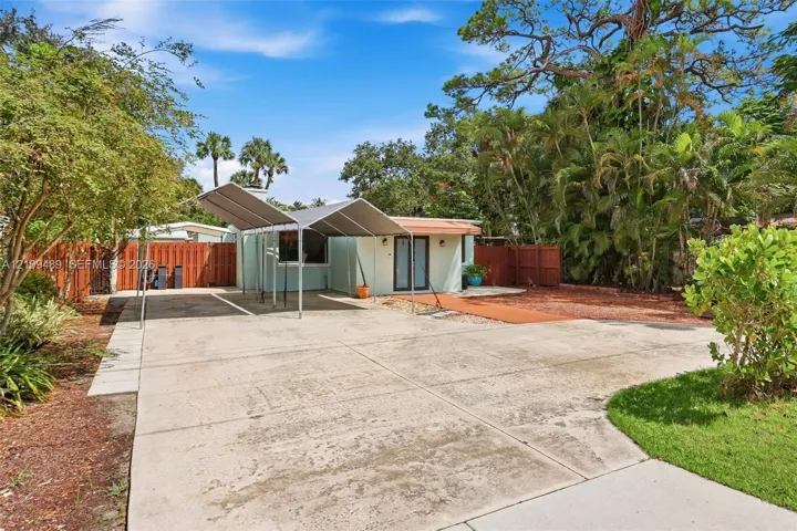 Exterior space featuring a concrete driveway, metal-framed carport, fenced perimeter, and mature landscape
