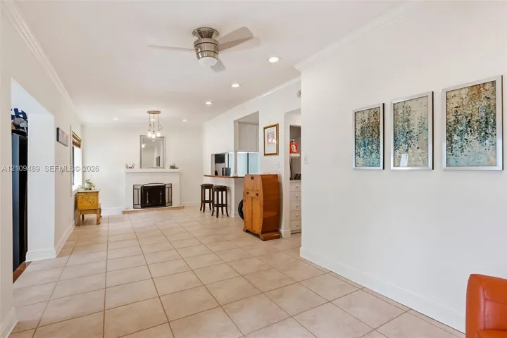Open interior space featuring light-toned tile flooring, recessed lighting, a ceiling fan, crown molding, and a fireplace with a mantel