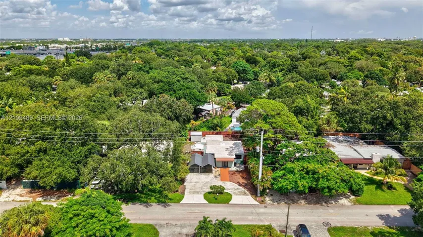 Aerial perspective featuring a single-story residence with a paved driveway, metal-framed carport, and mature tree canopy throughout the lot
