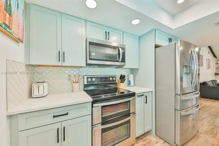 Kitchen featuring light blue cabinetry, stainless steel appliances, quartz countertops, arabesque-pattern tile backsplash, and wood-finish flooring