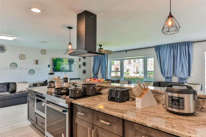 Kitchen featuring stone countertops, dark-finish cabinetry, an integrated cooktop with a stainless steel chimney-style range hood, tile flooring, and geometric pendant lighting
