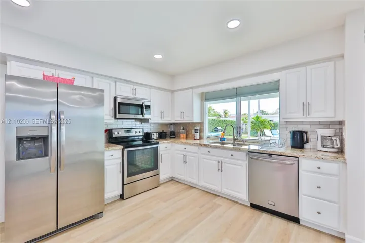 Kitchen featuring white shaker-style cabinetry, light-toned stone countertops, subway tile backsplash, wood-finish flooring, and stainless steel appliances