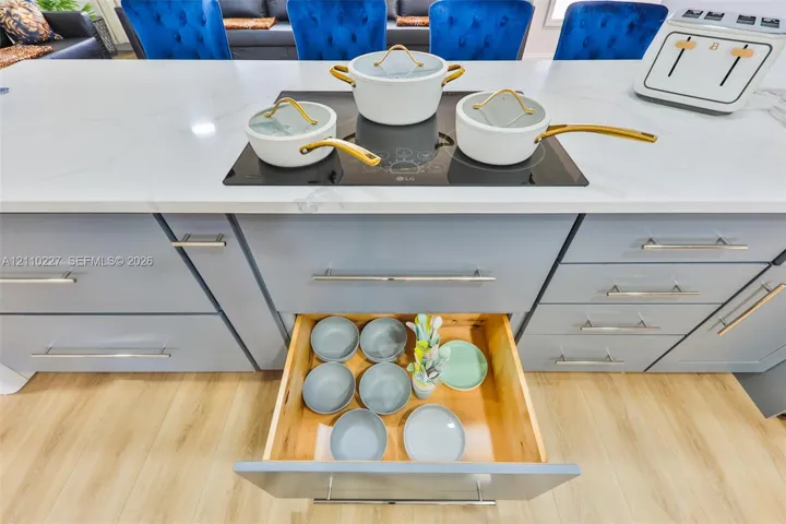 Kitchen island featuring a white surface with grey veining, a flush-mounted induction cooktop, light-grey flat-panel cabinetry with metal hardware, and deep-drawer storage with wood interiors, complemented by wood-finish flooring