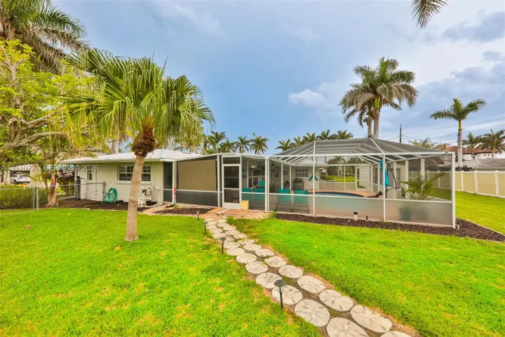Expansive backyard featuring a manicured lawn, stone paver pathway, mature palm trees, and an enclosed swimming pool with a screened lanai