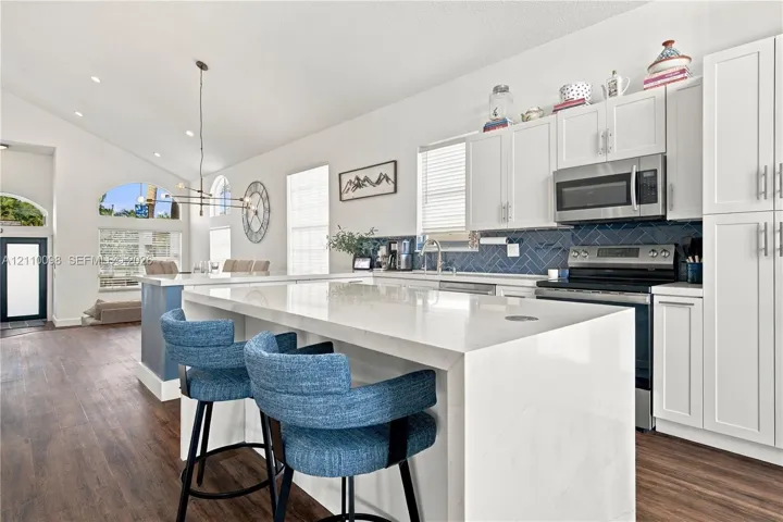 Kitchen featuring a quartz-topped island with waterfall edge, white shaker-style cabinetry, blue herringbone-pattern tile backsplash, stainless steel appliances, and wood-finish flooring