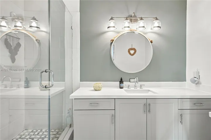 Bathroom featuring a vanity with light gray cabinetry, white solid-surface countertop, rectangular undermount sink, four-light vanity fixture, and a glass-enclosed shower with patterned tile flooring