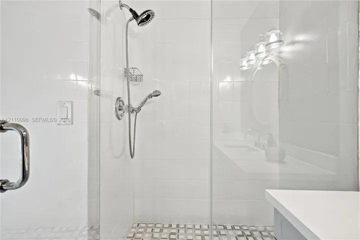Walk-in glass shower enclosure featuring white tile walls, a handheld showerhead, built-in wire shelving, and a patterned tile​​‌​​​​‌​​‌‌​‌‌​​​‌‌​‌​‌​‌​​​‌​​ floor