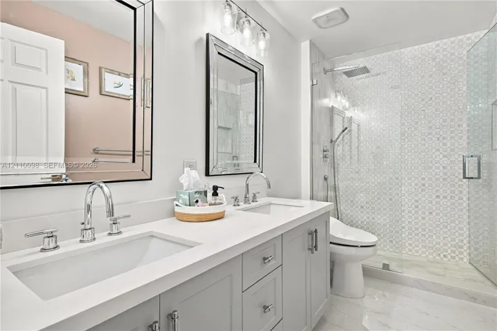 Dual-vanity cabinetry with a light-toned solid surface countertop, undermount sinks, polished chrome hardware, walk-in glass shower with decorative wall tile, and framed wall mirrors
