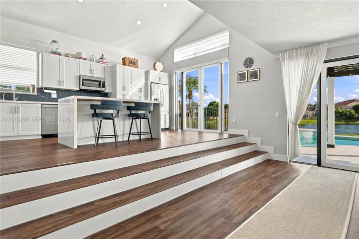 Kitchen featuring wood-finish flooring, white cabinetry, a center island with a waterfall countertop, vaulted ceilings, and glass-paned doors opening to an exterior patio