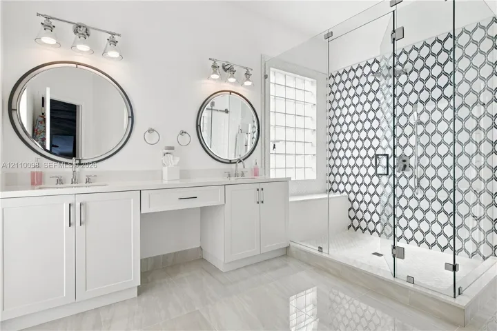 Double vanity featuring white cabinetry, oval mirrors, dual three-light fixtures, and a walk-in glass-enclosed shower with patterned tile surround