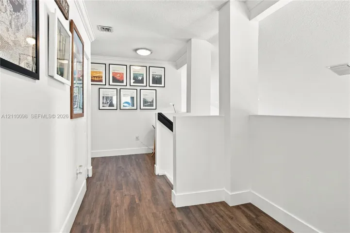Hallway featuring wood-finish flooring, crisp white walls, crown molding, and a staircase with a solid-surfaced wall and a contrasting handrail