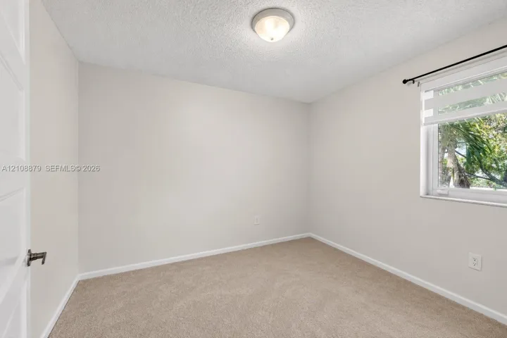 Neutral-toned interior space featuring wall-to-wall carpeting, a single-pane window with adjustable blinds, and a central flush-mount ceiling light fixture