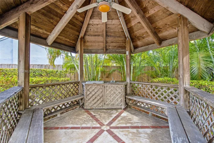 Covered gazebo featuring a wood-plank ceiling, structural timber posts, built-in wooden bench seating, a central ceiling fan, and an integrated outdoor prep station with a sink