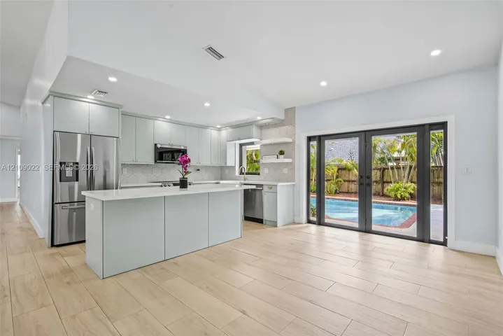 Kitchen featuring light-toned cabinetry, a central island with quartz countertops, stainless steel appliances, geometric tile backsplash, and wood-finish flooring with exterior access to an in-ground pool