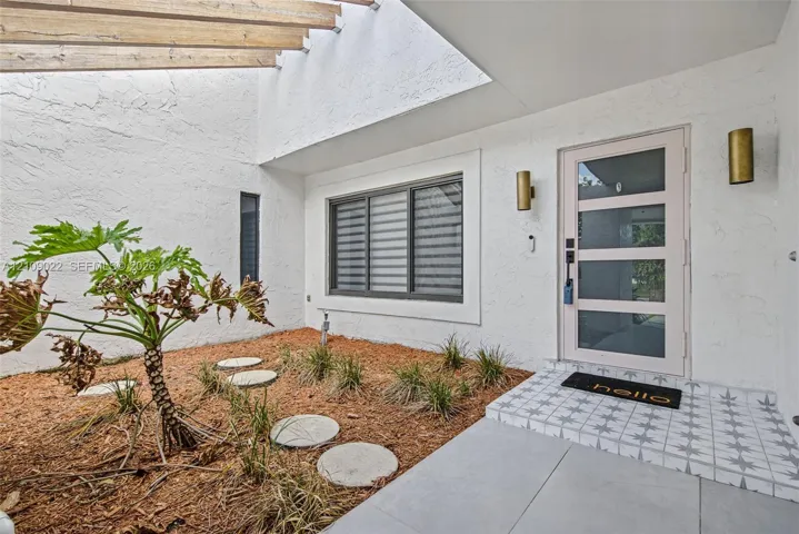 Entry courtyard featuring a timber beam pergola, textured stucco walls, decorative patterned tile stoop, and a contemporary glass-panel front door with brass wall-mounted sconces