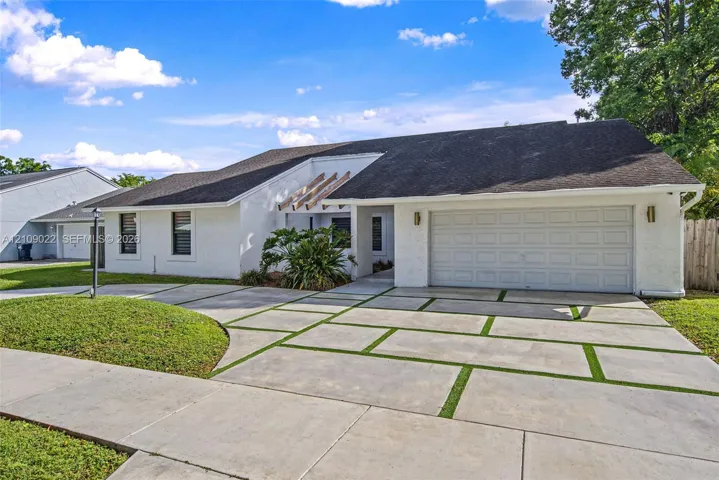 Single-story residence featuring a white exterior, multi-paneled garage door, geometric concrete paver driveway with integrated grass ribbons, and decorative wood roofline accents