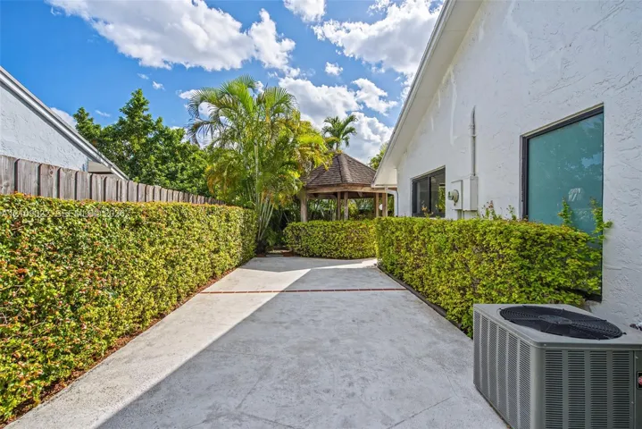 Concrete side yard featuring mature privacy hedging, a wooden gazebo, and textured exterior siding
