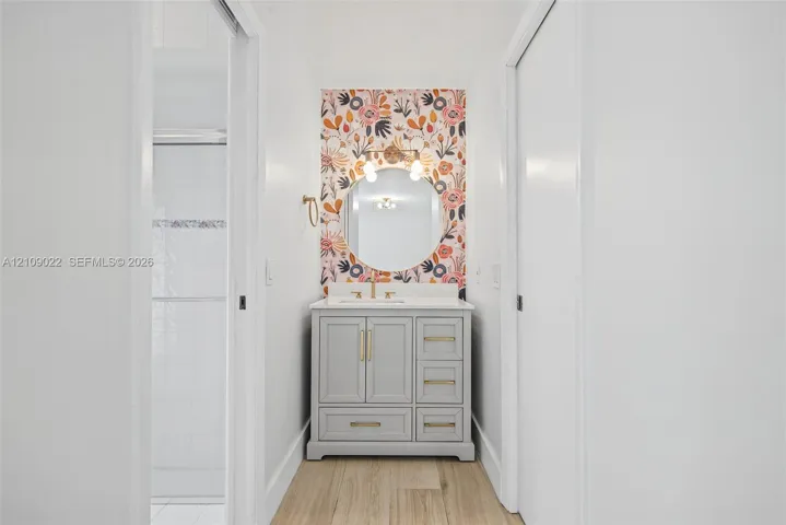 Gray vanity featuring a solid surface countertop, circular wall-mounted mirror, floral patterned wall accent, and metal cabinet hardware
