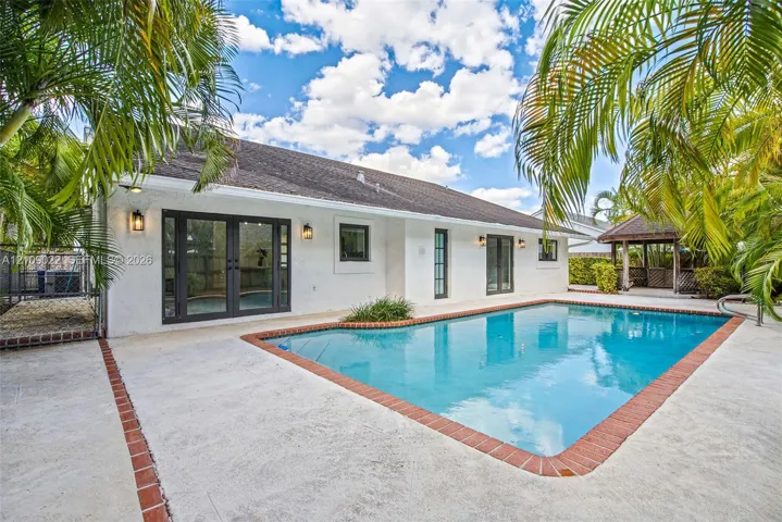In-ground swimming pool featuring a brick-tiled perimeter, concrete patio surround, exterior glass paneled double doors, and a detached gazebo