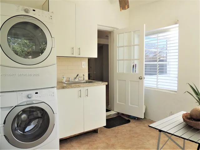 Utility area featuring white cabinetry, a stone-finish countertop with an integrated sink, stacked laundry appliances, tile flooring, and a multi-pane glass door