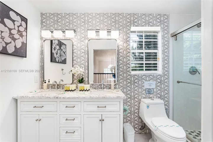 Dual-sink vanity with white cabinetry, patterned tile feature wall, rectangular mirrors, and a glass-enclosed shower