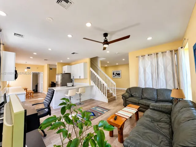 Open-concept interior featuring wood-finish flooring, recessed lighting, a modern kitchen with white cabinetry, a breakfast bar, and a staircase with a white railing