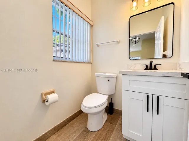 White shaker-style vanity with stone-top surface, matte black faucet, rectangular mirror with rounded corners, and wood-finish flooring