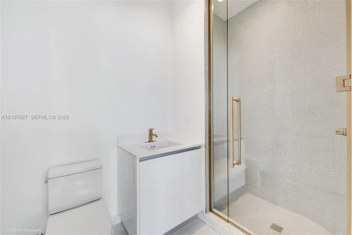 Modern bathroom featuring a glass-enclosed shower with small-format tile surround, a minimalist vanity with a white countertop and gold-tone faucet, and a sleek, low-profile toilet