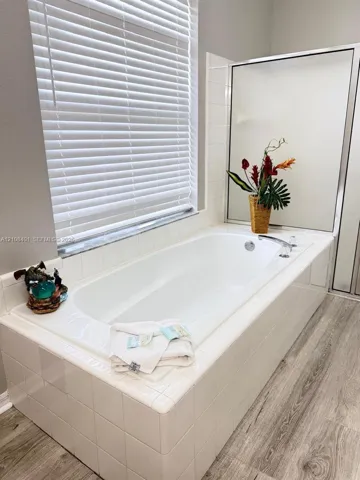 Built-in soaking tub with white tile surround, wood-finish flooring, and horizontal window blinds