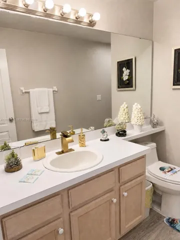 Bathroom vanity featuring a white countertop, inset oval sink, gold-toned faucet, wood-finish cabinetry, and a full-width wall mirror with integrated lighting