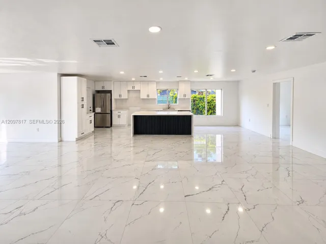 Open-concept space featuring white stone-finish tile flooring, recessed lighting, and a kitchen equipped with white cabinetry, a contrasting dark kitchen island, and stainless steel appliances