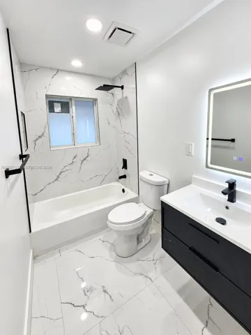 Bathroom featuring large-format stone-patterned tile flooring and wall surrounds, dark finish floating vanity with white countertop, matte black fixtures, and a wall-mounted LED mirror