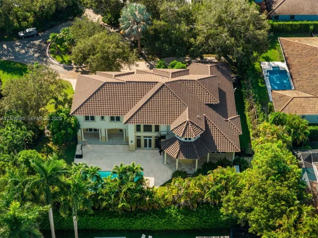 Aerial view of a two-story residence featuring a tile roof, light-toned exterior finish, stone-paved patio, private swimming pool, and dense perimeter landscaping