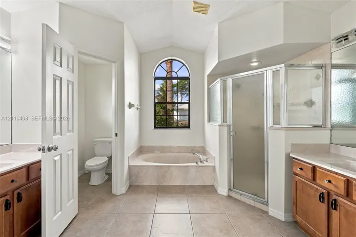 Bathroom featuring an arched window, a tiled soaking tub with a matching surround, a glass-enclosed shower, dual wood-finish vanities, and ceramic tile flooring