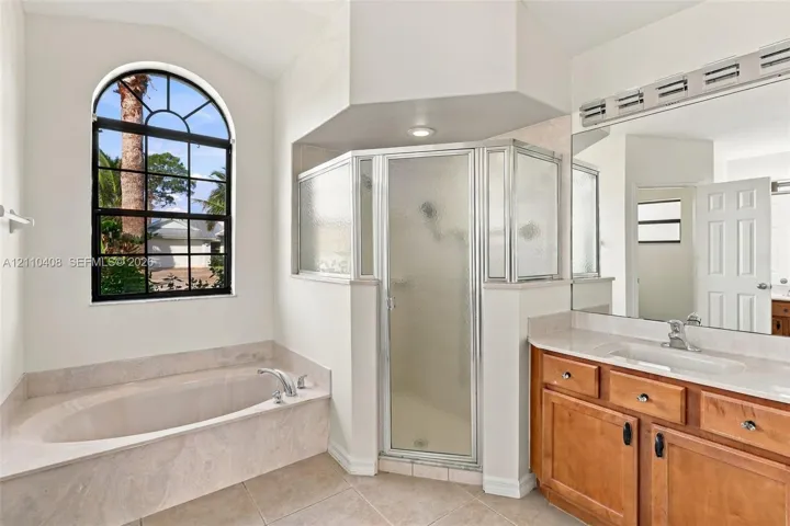 Arched window with exterior greenery, built-in soaking tub with stone-finish surround, glass-enclosed corner shower, wood-finish cabinetry, and vanity with stone-finish countertop