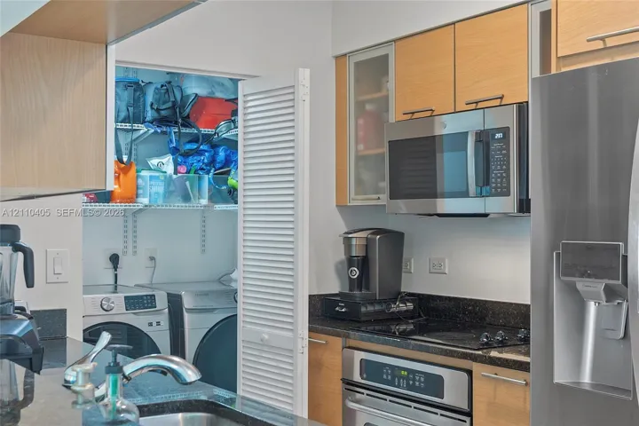 Kitchen featuring light-finish cabinetry, dark stone-style countertops, stainless steel appliances, and a closet with a louvered door providing integrated laundry space
