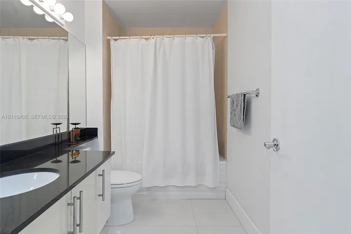 Bathroom featuring a vanity with dark stone countertop, integrated sink, polished metal hardware, full-width mirror with integrated lighting, and a bathtub with tile surround