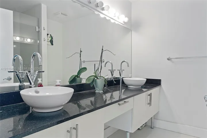 Dual vanity featuring a dark countertop, white high-gloss cabinetry, vessel sinks with polished chrome faucets, and a full-width mirror with integrated lighting